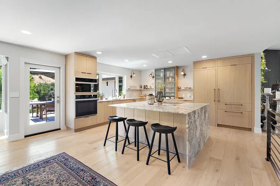 Elysium kitchen with marble island, black bar stools, built-in ovens, and patio door to hillside